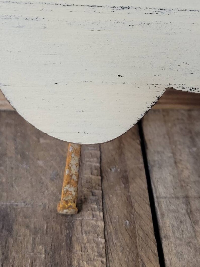 Puede incluir: Primer plano de un peque&ntilde;o objeto de madera blanquecina con un borde curvo y una sola pata de metal oxidado. La madera tiene un aspecto envejecido y desgastado con vetas visibles y detalles negros. La pata es rectangular y muestra signos de &oacute;xido.