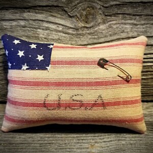 May include: A small, rectangular pillow with a red and white striped fabric and a blue and white American flag patch. The pillow has the word "USA" stitched on it and a safety pin is attached to the fabric.