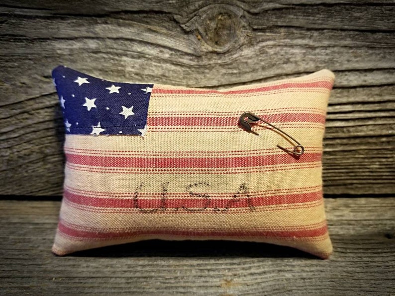 May include: A small, rectangular pillow with a red and white striped fabric and a blue and white American flag patch. The pillow has the word "USA" stitched on it and a safety pin is attached to the fabric.