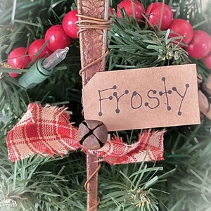 May include: A rustic Christmas ornament made from a vintage spoon, wrapped in wire, and decorated with a red and white plaid ribbon, a bell, and a brown paper tag that says "Frosty".