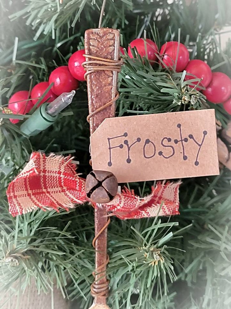 May include: A rustic Christmas ornament made from a vintage spoon, wrapped in wire, and decorated with a red and white plaid ribbon, a bell, and a brown paper tag that says "Frosty".