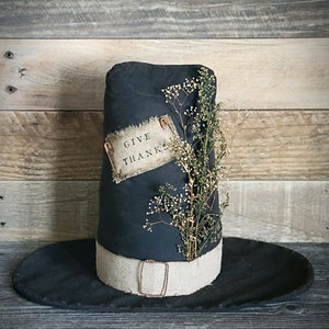 May include: A black pilgrim hat with a brown fabric band and a small sign that says "Give Thanks" attached to the side. Dried flowers are tucked into the hat.