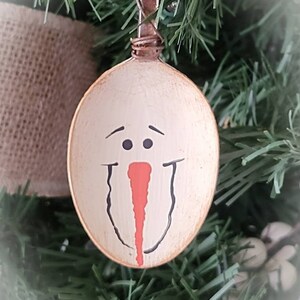 May include: A white painted wooden spoon ornament with a snowman face painted on it. The snowman has black eyes, a red nose, and a black mouth.