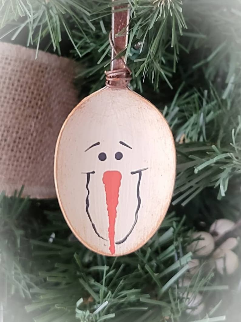 May include: A white painted wooden spoon ornament with a snowman face painted on it. The snowman has black eyes, a red nose, and a black mouth.