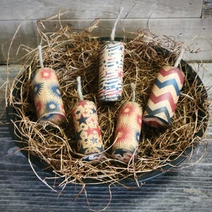 May include: A collection of six decorative firecracker-shaped items with patriotic designs. Each is cylindrical, wrapped in paper with red, white, and blue patterns, and has a white string fuse. They are arranged in a black bowl filled with straw.