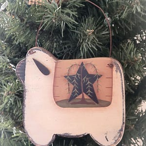 Puede incluir: Un adorno de madera blanco con forma de oveja con una estrella negra y un árbol verde pintados en la parte posterior. El adorno tiene un colgador de alambre.