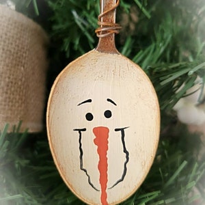 May include: A white painted metal spoon with a snowman face painted on it. The snowman has black eyes, a black mouth, and a red nose.