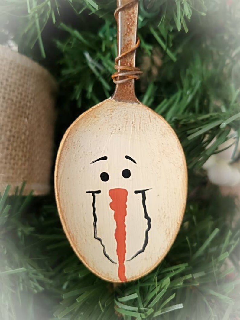May include: A white painted metal spoon with a snowman face painted on it. The snowman has black eyes, a black mouth, and a red nose.