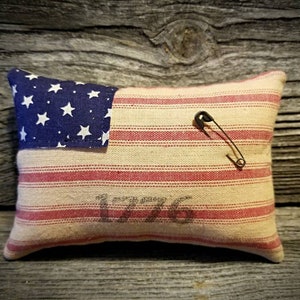 May include: A small pillow shaped like the American flag with red and white stripes and a blue field of stars. The pillow has a safety pin attached to it and the year 1776 printed on the front.