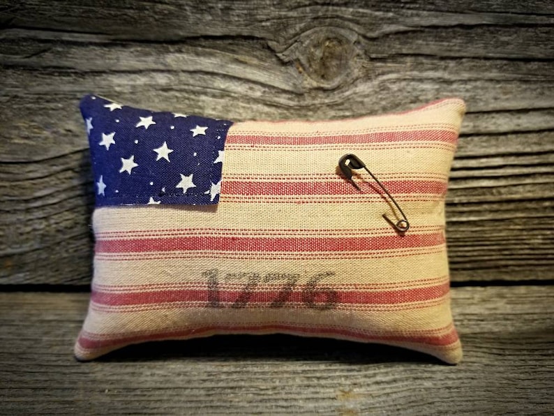 May include: A small pillow shaped like the American flag with red and white stripes and a blue field of stars. The pillow has a safety pin attached to it and the year 1776 printed on the front.