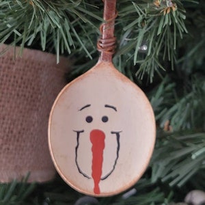 May include: A white painted wooden spoon ornament with a snowman face painted on it. The snowman has black eyes, a red nose, and a black smile.