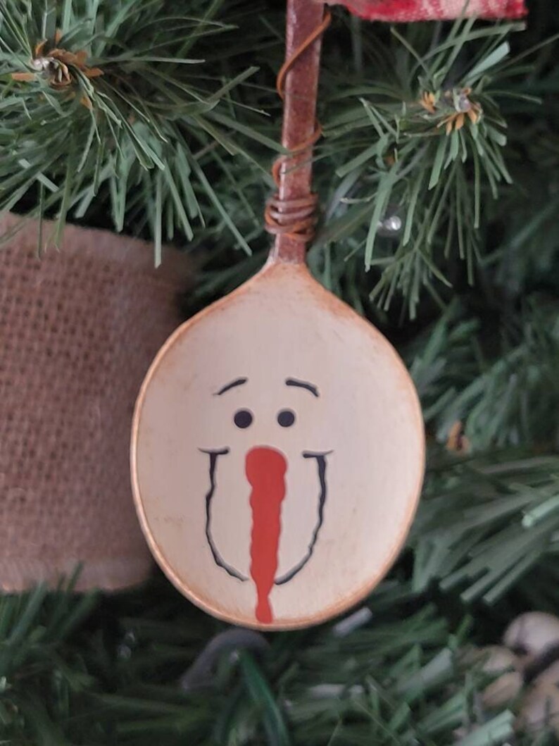 May include: A white painted wooden spoon ornament with a snowman face painted on it. The snowman has black eyes, a red nose, and a black smile.