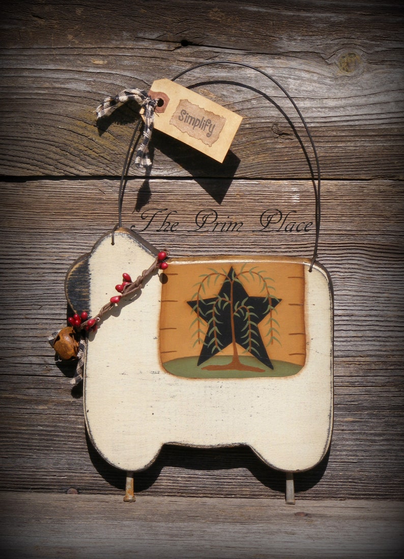 Puede incluir: Adorno de pared con forma de oveja de color crema con un dise&ntilde;o de estrella y &aacute;rbol negro. Cuenta con una etiqueta que dice "Simplify" y est&aacute; adornado con una peque&ntilde;a campana y bayas. "The Prim Place" est&aacute; escrito en la parte superior.