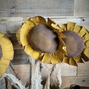 May include: Three rustic-style felt sunflowers with brown centers and yellow petals, arranged in a vase with burlap fabric. The sunflowers are on a wooden background.