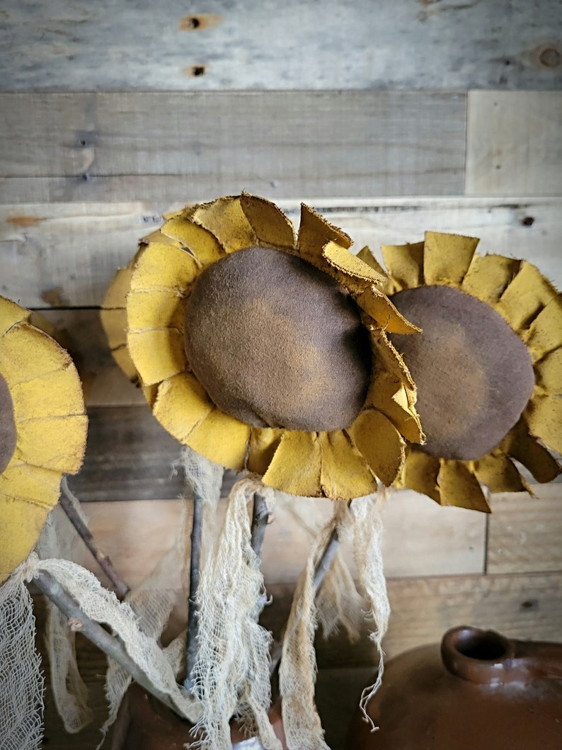 May include: Three rustic-style felt sunflowers with brown centers and yellow petals, arranged in a vase with burlap fabric. The sunflowers are on a wooden background.