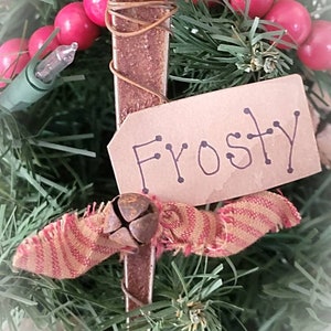 May include: A rustic Christmas ornament made from a rusty metal piece with a red and gold striped fabric bow and a small bell. The ornament has a brown paper tag with the word "Frosty" written on it.