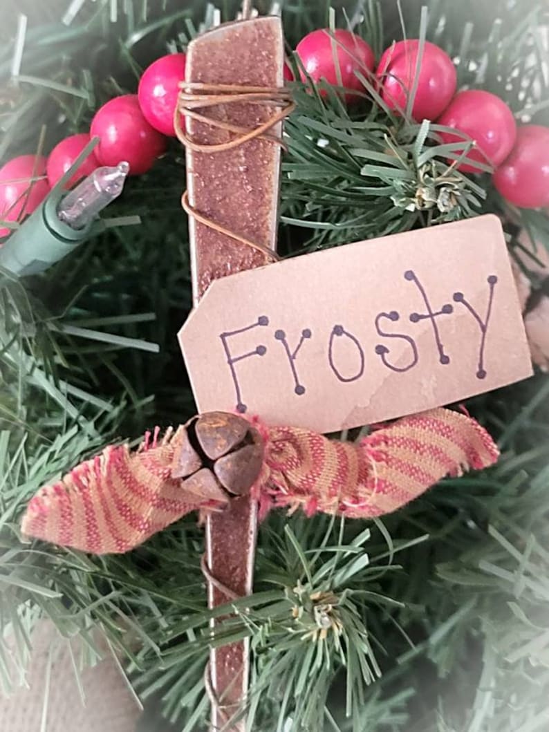 May include: A rustic Christmas ornament made from a rusty metal piece with a red and gold striped fabric bow and a small bell. The ornament has a brown paper tag with the word "Frosty" written on it.