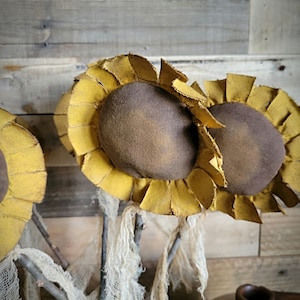 May include: Three rustic-style felt sunflowers with brown centers and yellow petals, arranged in a vase with burlap fabric. The sunflowers are on a wooden background.
