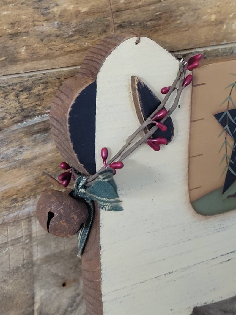 Puede incluir: Adorno navide&ntilde;o de madera con forma de reno, con una campana r&uacute;stica, una ramita de bayas rojas y una cinta a cuadros verde. El reno tiene orejas pintadas de negro y un cuerpo color crema. El adorno est&aacute; dise&ntilde;ado para colgar.