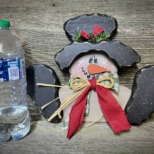May include: A wooden snowman ornament with a black hat, red scarf, and a brown bow. The snowman has a white face with black eyes, a carrot nose, and a black mouth.