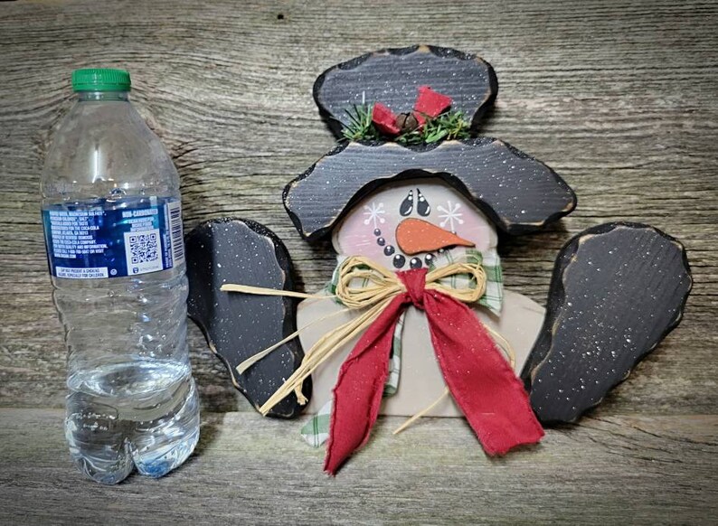 May include: A wooden snowman ornament with a black hat, red scarf, and a brown bow. The snowman has a white face with black eyes, a carrot nose, and a black mouth.