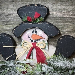 May include: A wooden snowman ornament with a black top hat, black arms, and a white face with a red scarf and a brown bow. The snowman is surrounded by green pine branches with white snow.