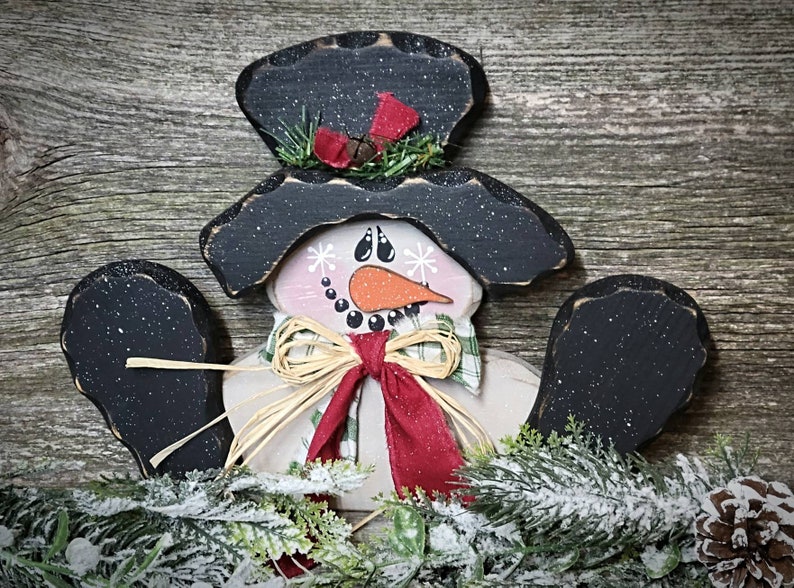 May include: A wooden snowman ornament with a black top hat, black arms, and a white face with a red scarf and a brown bow. The snowman is surrounded by green pine branches with white snow.
