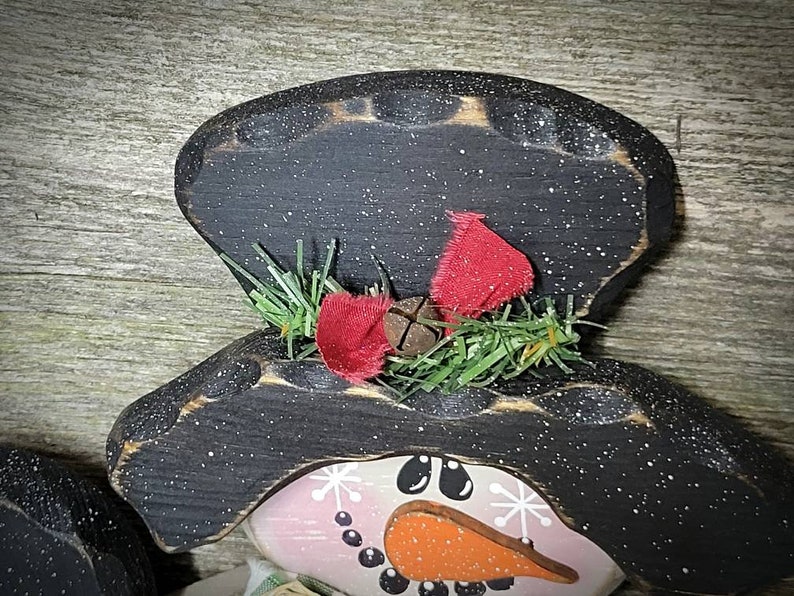May include: A wooden snowman ornament with a black hat, red bow, and green pine sprigs. The snowman's face is painted with black eyes, an orange nose, and a pink mouth.