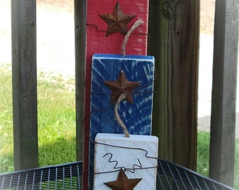 May include: Three wooden blocks painted red, white, and blue, each with a rusty metal star and twine loop. The blocks are arranged vertically, evoking a patriotic theme. The blocks are on a metal grate.