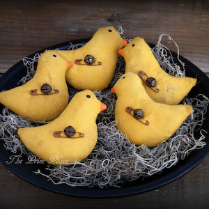 May include: Five yellow felt bird ornaments with orange beaks and brown safety pin accents. The birds are arranged in a nest of brown and gray straw-like material on a black plate.