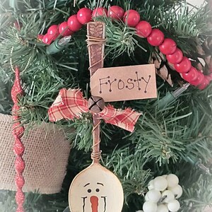 May include: A spoon ornament painted to look like a snowman with a red nose and black eyes. The spoon is attached to a red and white plaid ribbon and a brown tag that says "Frosty".