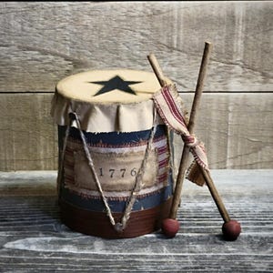 May include: A decorative drum with a black star on top and the year "1776" on the side. The drum is made of wood and fabric, with drumsticks resting against it. The color scheme includes red, blue, and beige.