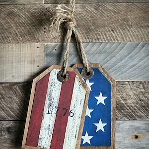 Puede incluir: Dos etiquetas de madera con un acabado de pintura desgastado. Una etiqueta es roja, blanca y azul con el número 1776. La otra etiqueta es azul con estrellas blancas. Las etiquetas están atadas juntas con un cordel.