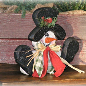 May include: A wooden snowman figurine with a black top hat, red scarf, and plaid bow. The snowman is sitting on a wooden surface with a rustic background.