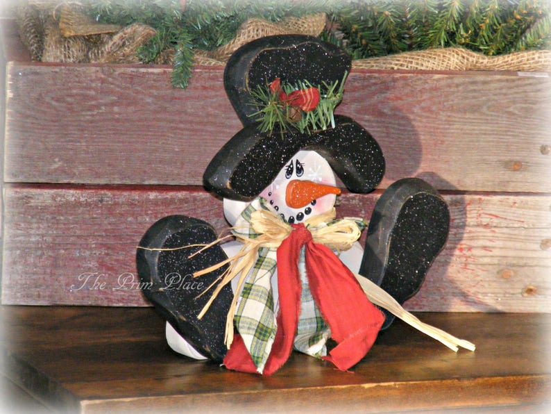 May include: A wooden snowman figurine with a black top hat, red scarf, and plaid bow. The snowman is sitting on a wooden surface with a rustic background.