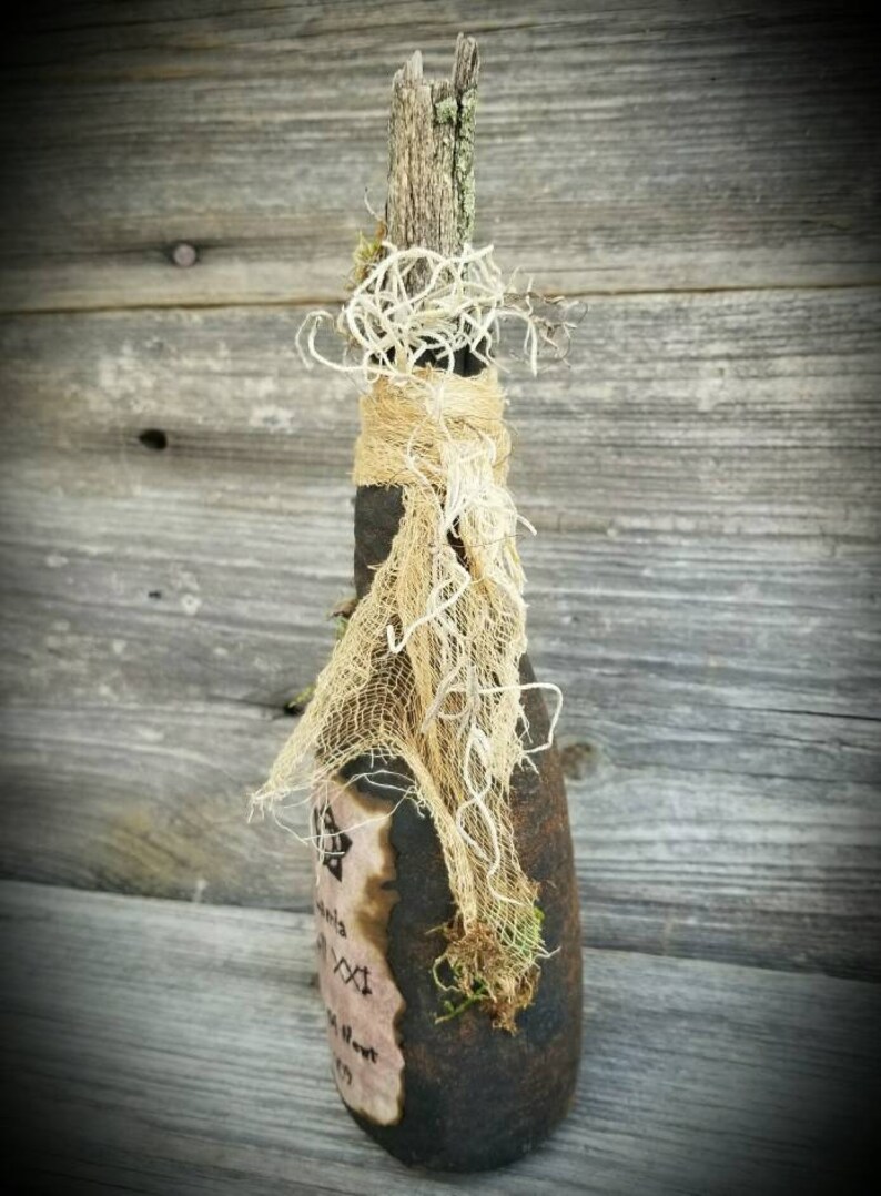 Op de afbeelding: Een donkerbruine glazen fles met een rustieke, verweerde uitstraling. Hij is versierd met een stuk jute stof, mos en een takje. De fles heeft een etiket met handgeschreven tekst en een stervormig ontwerp.