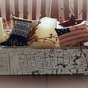 May include: A rustic wooden box centerpiece decorated with red, white, and blue patriotic themed fabric, candles, and sprigs of berries. The box is painted white with a distressed finish and has the word "liberty" written on it.