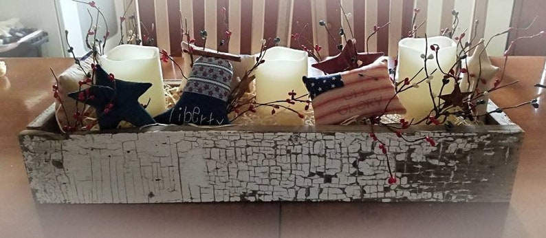 May include: A rustic wooden box centerpiece decorated with red, white, and blue patriotic themed fabric, candles, and sprigs of berries. The box is painted white with a distressed finish and has the word "liberty" written on it.