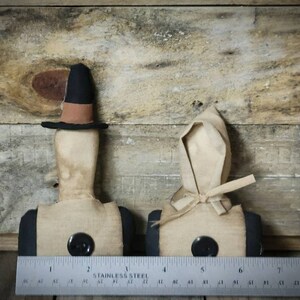 May include: Two primitive fabric dolls with black buttons and simple features. The doll on the left is wearing a black hat with a brown band. The doll on the right is wearing a white bonnet. Both dolls are standing on a wooden surface.