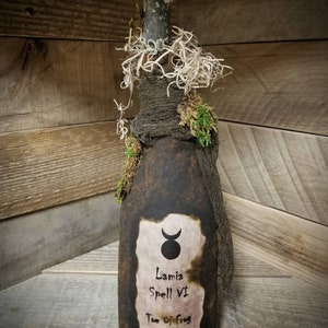 Op de afbeelding: Een donkerbruine glazen fles met een kurkstop, gewikkeld in jute en mos. De fles heeft een etiket met een halve maan en een symbool, en de tekst "Lamia Spell VI Toe of Frog 1609".