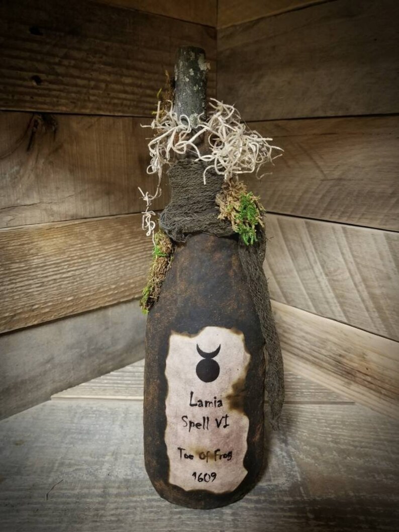 Op de afbeelding: Een donkerbruine glazen fles met een kurkstop, gewikkeld in jute en mos. De fles heeft een etiket met een halve maan en een symbool, en de tekst "Lamia Spell VI Toe of Frog 1609".