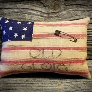 May include: A small pillow with a red, white, and blue striped fabric and a blue fabric star patch. The pillow has the words "Old Glory" stitched on it. A safety pin is attached to the pillow.