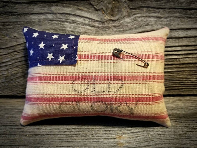 May include: A small pillow with a red, white, and blue striped fabric and a blue fabric star patch. The pillow has the words "Old Glory" stitched on it. A safety pin is attached to the pillow.