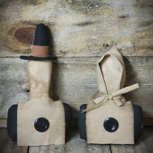 May include: Two primitive style fabric dolls with black buttons for eyes. The doll on the left is wearing a black hat with a brown band. The doll on the right is wearing a white bonnet. Both dolls are standing on a wooden surface.