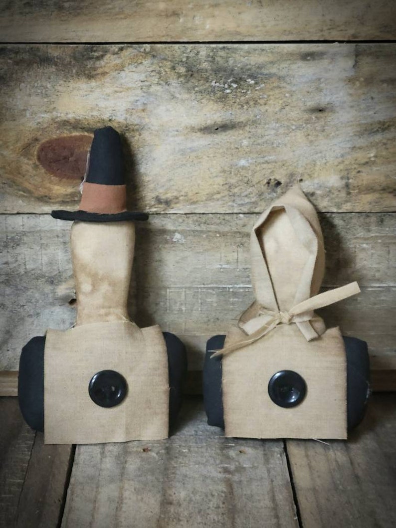 May include: Two primitive style fabric dolls with black buttons for eyes. The doll on the left is wearing a black hat with a brown band. The doll on the right is wearing a white bonnet. Both dolls are standing on a wooden surface.