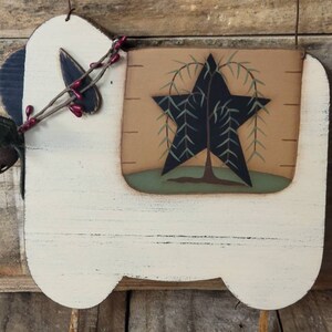Puede incluir: Adorno de pared r&uacute;stico de madera con forma de oveja. La oveja est&aacute; pintada de crema con una oreja azul oscuro y un dise&ntilde;o decorativo de estrella y &aacute;rbol. Cuenta con una peque&ntilde;a campana y una ramita de bayas. El fondo es de madera envejecida.
