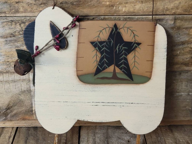 Puede incluir: Adorno de pared r&uacute;stico de madera con forma de oveja. La oveja est&aacute; pintada de crema con una oreja azul oscuro y un dise&ntilde;o decorativo de estrella y &aacute;rbol. Cuenta con una peque&ntilde;a campana y una ramita de bayas. El fondo es de madera envejecida.