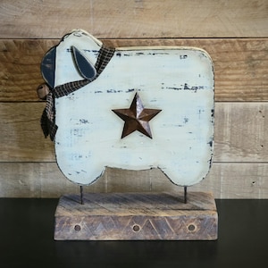 May include: A rustic, distressed wooden sheep figurine with a cream-colored body and a brown star. The sheep has a black and brown scarf and ears. It stands on a wooden base, perfect for farmhouse decor.