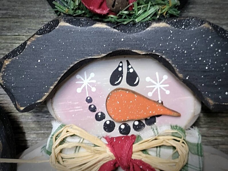 May include: A wooden snowman ornament with a black hat, a pink face, a carrot nose, and black eyes. The snowman is wearing a green and white plaid scarf with a brown straw bow.