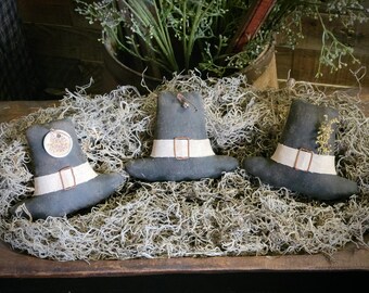 May include: Three dark gray fabric pilgrim hats with cream-colored bands and copper-colored buckles. The left hat features a round tag with the text Give Thanks. The right hat is adorned with a small sprig of dried yellow flowers.
